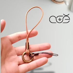 Könnte beinhalten: Ein Handy-Anhänger aus hellbraunem Leder mit einem bronzefarbenen Metallring und Verschluss. Der Anhänger hat eine Schlaufe für das Handgelenk und eine kleine schwarze Kordel. Das Logo "aöe" ist in Schwarz sichtbar.