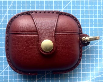 May include: A burgundy leather AirPod case with a brass button closure and a gold-colored clasp. The case has a stitched border and a vertical strap detail. The case is designed to protect and carry wireless earbuds.