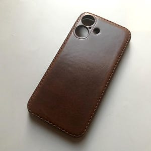 Puede incluir: Una funda de teléfono de cuero marrón con un borde cosido y un recorte para el objetivo de la cámara.