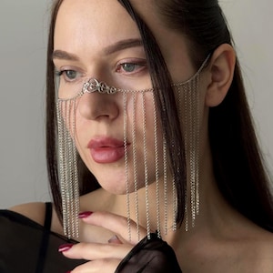 May include: Silver face jewellery featuring a delicate chain design that drapes over the eyes and nose. The face chain has a decorative element above the nose and long, dangling chains. The jewellery is worn on the face.