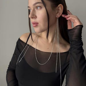 Op de afbeelding: Zilveren kettingoorbellen met lange, bungelende strengen. De oorbellen worden gedragen met een zwarte top. Het model kijkt opzij. De oorbellen hebben een eenvoudig, elegant ontwerp.