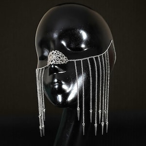 Puede incluir: Joya facial plateada con una pieza nasal de filigrana decorativa y cadenas colgantes con extremos puntiagudos. Las cadenas caen en cascada por la cara, creando un efecto dramático. Este accesorio único está diseñado para ser usado en la cara.