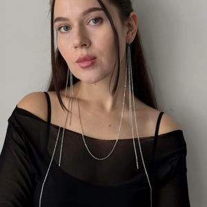 May include: Silver chain earrings and necklace. The earrings are long, dangling chains. The necklace is a simple chain. The woman is wearing a black top.