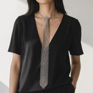 May include: A silver chainmail tie necklace. The necklace is worn over a black V-neck top and black trousers. The tie is made of small, interconnected metal rings, creating a textured, metallic appearance. The model is standing in front of a white background.