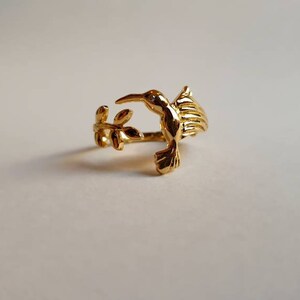 Può includere: Un anello dorato con un colibrì in volo, posato su un ramo con foglie. L'anello è in metallo e ha un design delicato.