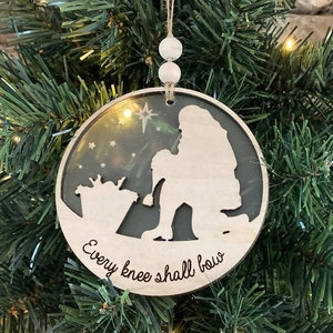 May include: A wooden Christmas ornament with a silhouette of a person kneeling and the text "Every knee shall bow".