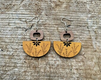May include: A pair of wooden earrings with a floral design. The earrings are made of two pieces of wood, one a half-circle and the other a rectangle with a cut-out. The half-circle piece has a floral design etched into it, and the rectangle piece has a small flower design in the center.
