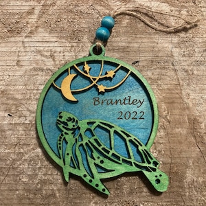 May include: A teal and green wooden ornament shaped like a sea turtle with a crescent moon and stars in the background. The ornament is personalized with the name "Brantley" and the year "2022".