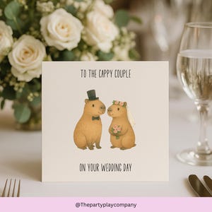 Könnte beinhalten: Quadratische Hochzeitskarte mit einer Illustration eines Capybara-Paares. Das Capybara links trägt einen Zylinder und eine Fliege, das andere einen Blumenkranz und hält einen Blumenstrauß. Text: "To the Cappy Couple On Your Wedding Day".