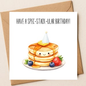 May include: A birthday card featuring a stack of pancakes with a pat of butter and syrup, a party hat, and a smiling face. The card includes the text "HAVE A SPEC-STACK-ULAR BIRTHDAY!" with a strawberry and blueberries.