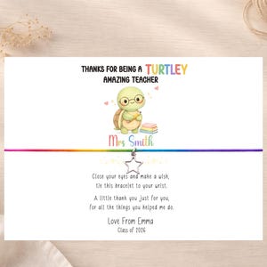Könnte beinhalten: Eine weiße Karte mit einem Regenbogen-Armband. Die Karte zeigt eine Zeichentrickschildkröte mit Brille und Büchern. Der Text lautet "Danke, dass Sie eine tolle Lehrerin sind" und "Von Emma, Klasse 2026".