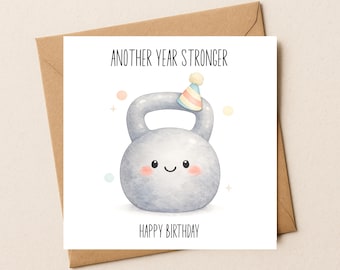 May include: A square greeting card featuring a watercolor illustration of a grey kettlebell with a smiling face, pink cheeks, and a small striped party hat. The text reads ANOTHER YEAR STRONGER at the top and HAPPY BIRTHDAY at the bottom.