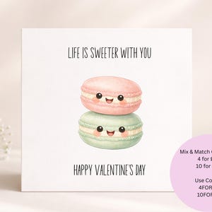 Könnte beinhalten: Quadratische Grußkarte mit dem Text "LIFE IS SWEETER WITH YOU" und "HAPPY VALENTINE'S DAY". Die Karte zeigt zwei Cartoon-Makronen, einen rosa und einen grünen, übereinander gestapelt, jeweils mit einem lächelnden Gesicht.