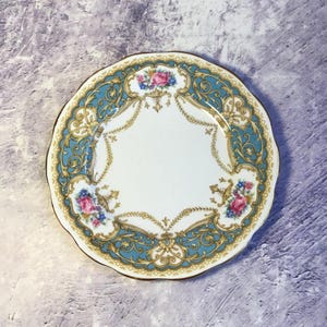 Puede incluir: Un plato de porcelana blanca con un diseño floral azul y dorado. El plato tiene un borde festoneado y presenta un delicado patrón floral con flores rosas y amarillas.