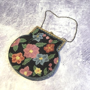 Puede incluir: Un bolso de aguja negro vintage con un diseño floral. El bolso presenta flores rosas, azules, amarillas y naranjas con hojas verdes. Tiene un marco de metal dorado y una cadena.