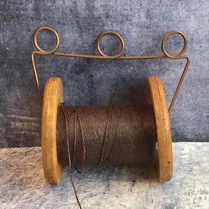 Puede incluir: Una bobina de hilo marrón de madera vintage con un soporte de hilo de alambre de latón. La bobina tiene extremos de madera y está llena de hilo bien enrollado. El alambre de latón tiene tres bucles circulares en la parte superior.
