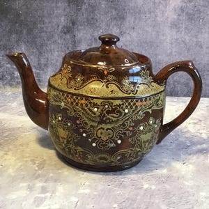 Könnte beinhalten: Eine braune Teekanne aus Keramik mit einem goldenen und orangefarbenen Blumendesign. Die Teekanne hat einen Deckel und einen Griff.