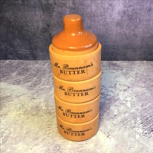May include: A vintage butter dish, stacked in sections, with a lid. The dish is light brown with the words "Mr. Brannam's BUTTER" printed in black. The lid is a darker brown. A classic kitchenware item.