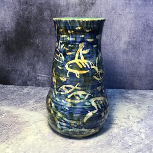 Peut inclure: Un vase en céramique avec un bord évasé et une base arrondie, présentant une surface émaillée bleu foncé et vert décorée de motifs de poissons jaunes peints à la main. L'extérieur affiche des textures de coups de pinceau horizontaux.