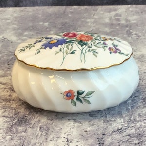May include: White porcelain trinket box with a floral design. The lid has a gold trim and features a variety of flowers in shades of pink, blue, yellow, and purple. The base of the box has a single flower design in shades of orange and blue.