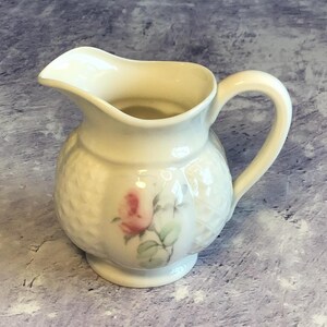 May include: Cream-coloured ceramic creamer with a rose design. The creamer has a rounded body with a textured pattern, a curved handle, and a spout. The rose is pink with green leaves. A decorative piece for serving.