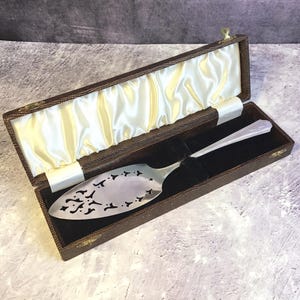 Puede incluir: Un servidor de pastel plateado con un diseño calado decorativo, presentado en una caja marrón texturizada forrada con satén y terciopelo negro. La caja es rectangular con un cierre dorado.