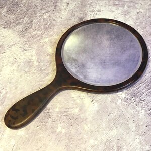 May include: A vintage-style hand mirror with a tortoiseshell pattern on the handle and frame. The mirror has a round shape and a reflective surface. The handle is curved for easy grip.