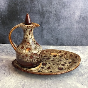 May include: A brown and cream ceramic jug with a spout and a handle, sitting on a matching brown and cream ceramic tray. The jug and tray have a speckled glaze.