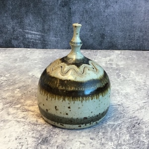 May include: A small, ceramic, brown and white, lidded jar with a rounded body and a tall, thin lid. The jar has a wavy design around the top and a brown stripe around the bottom.
