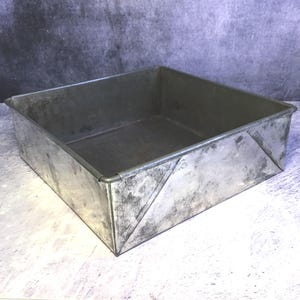 May include: A square, vintage metal baking tin with a distressed, weathered appearance. The tin has a rectangular shape with angled sides and a slightly raised rim. The metal has a silver-grey colour with darker areas, suggesting age and use. Ideal for baking cakes and other treats.