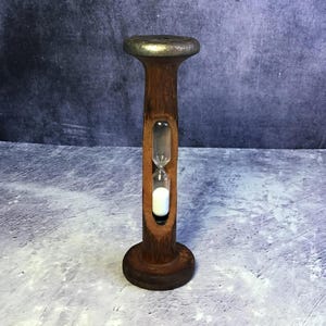 May include: A wooden hourglass with a dark brown finish and a silver-colored top. The hourglass contains white sand and is encased in a carved wooden frame. The base is round, and the overall design is vintage.