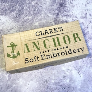 May include: A rectangular box of Clark's Anchor Soft Embroidery thread. The box is light brown with green text and an anchor graphic. The text reads "Anchor Fast Colour Soft Embroidery".