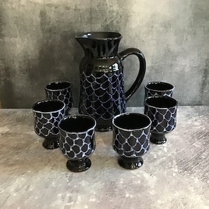 May include: A set of six black and white ceramic cups and a matching jug. The cups have a white geometric pattern on a black background. The jug has a handle and a spout.