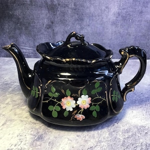 May include: Black ceramic teapot with a lid, spout, and handle. The teapot is decorated with pink and white floral designs and green leaves. Gold trim accents the edges and handle. A vintage tea set.