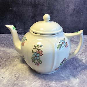 May include: A white teapot with a floral design featuring blue, pink, and green flowers. The teapot has a lid with a knob on top.