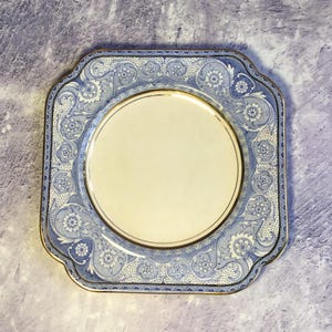 May include: A square, cream-colored plate with a blue and gold floral design. The plate has a raised edge and a scalloped rim.