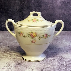 May include: Cream-coloured, lidded sugar bowl with two handles and a pedestal base. The bowl is decorated with a floral pattern of pink and yellow roses and gold trim. The lid has a small handle.
