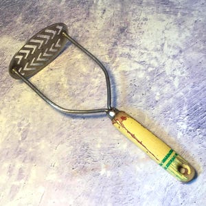 Pode incluir: Um espremedor de batatas de cozinha com uma cabeça de metal prateado com um padrão de corte em chevron repetitivo. A ferramenta tem uma estrutura de metal resistente ligada a um cabo cilíndrico de madeira pintado de amarelo com desgaste.