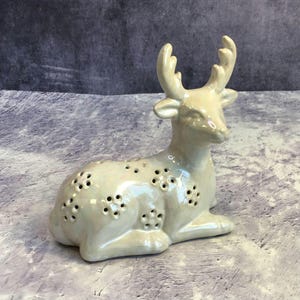 May include: A white ceramic deer figurine with a detailed design of small holes throughout the body. The deer is lying down with its head turned to the side.