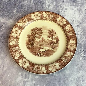 May include: A vintage brown and cream ceramic plate featuring a floral border and a scene of a countryside landscape with a castle, trees, and a cow.