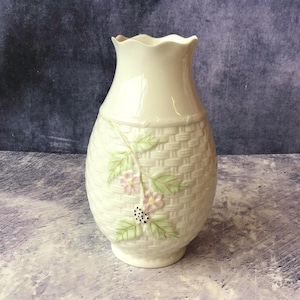 May include: Cream-coloured ceramic vase with a woven basket pattern on the lower half. It features a floral design with pink flowers, green leaves, and a cluster of black dots. The top has a scalloped rim.