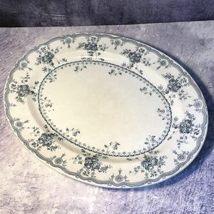 Peut inclure: Un plat de service ovale en céramique avec une base de couleur crème et un motif floral gris foncé. Le design comprend une bordure détaillée de roses et de filigranes complexes, avec une guirlande florale assortie entourant le centre.