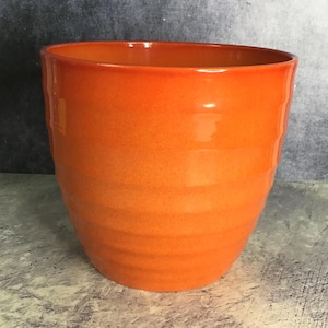 May include: A bright orange plant pot with a glossy finish and a series of horizontal, subtle ridges encircling the exterior. The vessel features a wide, open circular top and a tapered base, showcasing a vibrant, solid colour throughout the surface.