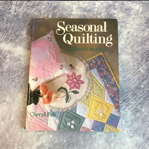 May include: The cover of a book, "Seasonal Quilting: A Year in Stitches", showcases a patchwork of quilting designs. These include floral patterns and decorative elements. The author's name, Cheryl Fall, is printed on the cover.