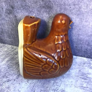 May include: A brown ceramic bird-shaped vase with a glossy finish. The bird has detailed wing and feather patterns. The vase has a light beige section on the side and a hole at the top for flowers. A decorative home decor item.