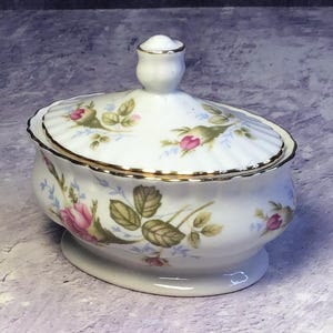 Puede incluir: Un azucarero de porcelana blanca con tapa, con un estampado floral de rosas rosas, verdes y azules. El borde y el asa están adornados con dorado. El cuenco tiene una base de pedestal.