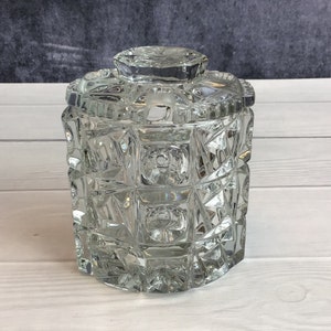 May include: A clear glass jar with a lid, featuring a geometric pattern of squares and triangles. The jar is made of thick, heavy glass and has a vintage look.