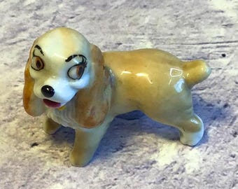 May include: A small ceramic figurine of Lady, the dog from Disney's Lady and the Tramp. The figurine is painted in shades of brown and white, with detailed facial features and a sweet expression.