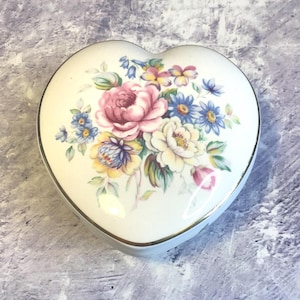 Puede incluir: Una caja de corazón blanca con un borde dorado y un diseño floral con flores rosas, amarillas y azules.