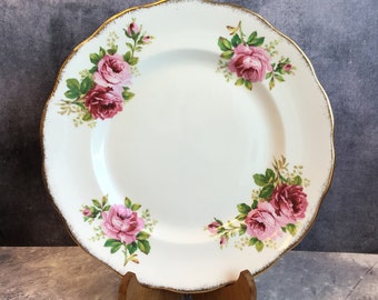 May include: A white ceramic dinner plate with a gold rim and a floral design featuring pink roses and green leaves.
